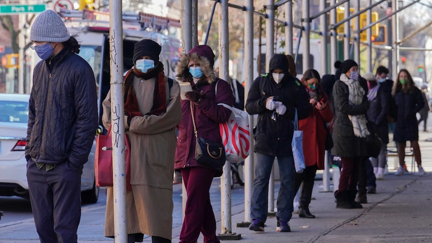 A line of people dressed in jackets, scarves and beanies wear masks as they stand on a street.