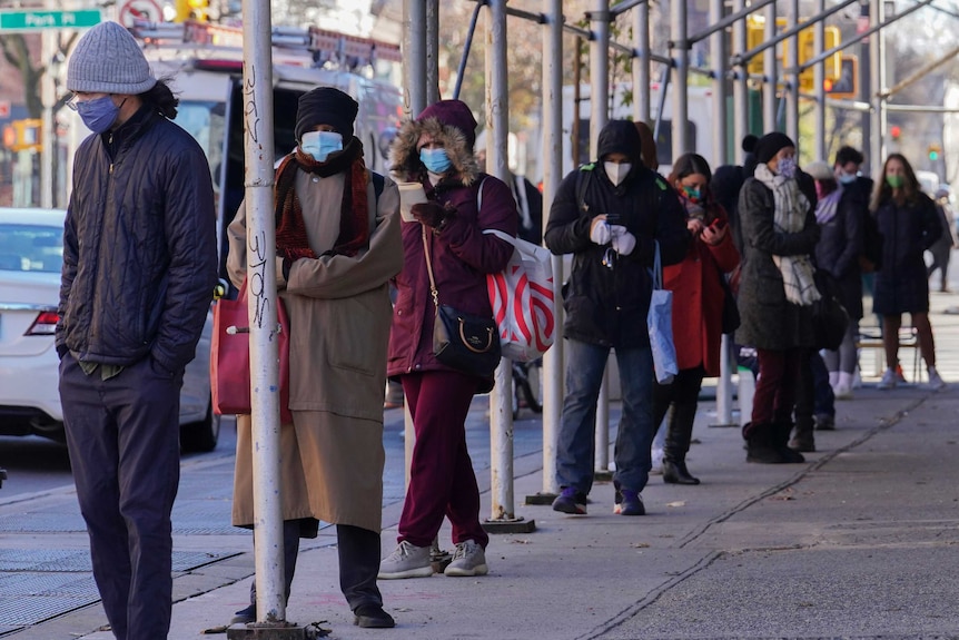 A line of people dressed in jackets, scarves and beanies wear masks as they stand on a street.