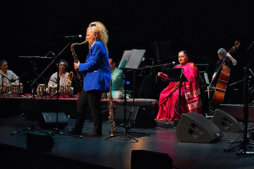 A woman plays saxophone on stage. Seated behind her is an Indian woman in a pink sari singing, beside other musicians.