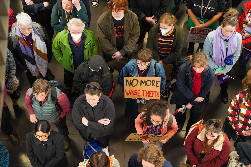 A crowd of protesters, mostly women, is seen from above, with a large "no more wage theft" sign in the middle.