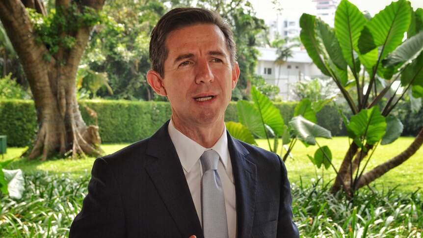 Simon Birmingham looks to the right of the camera as he stands in front of a lush garden. He wears a suit and is mid-speech.