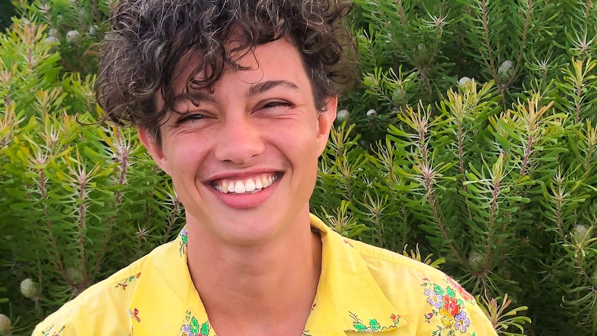 Rory Blundell smiles and looks to the camera while wearing a yellow shirt and standing in front of a bush.