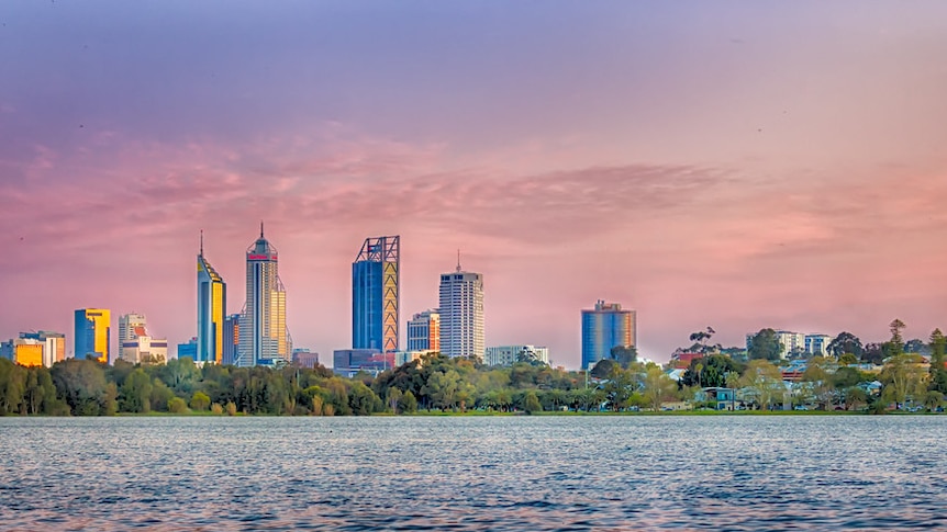 Calm water in the foreground reflecting a pink sunset. Tall buildings of Perth city centre are seen above trees on the foreshore