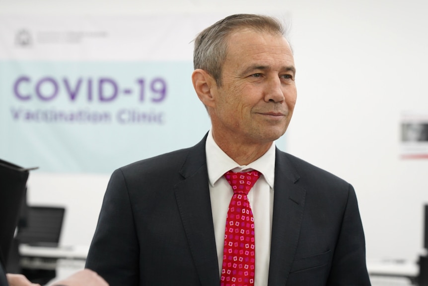 A mid-shot of WA Health Minister Roger Cook standing in front of a COVID-19 vaccination clinic sign, wearing a suit and tie.