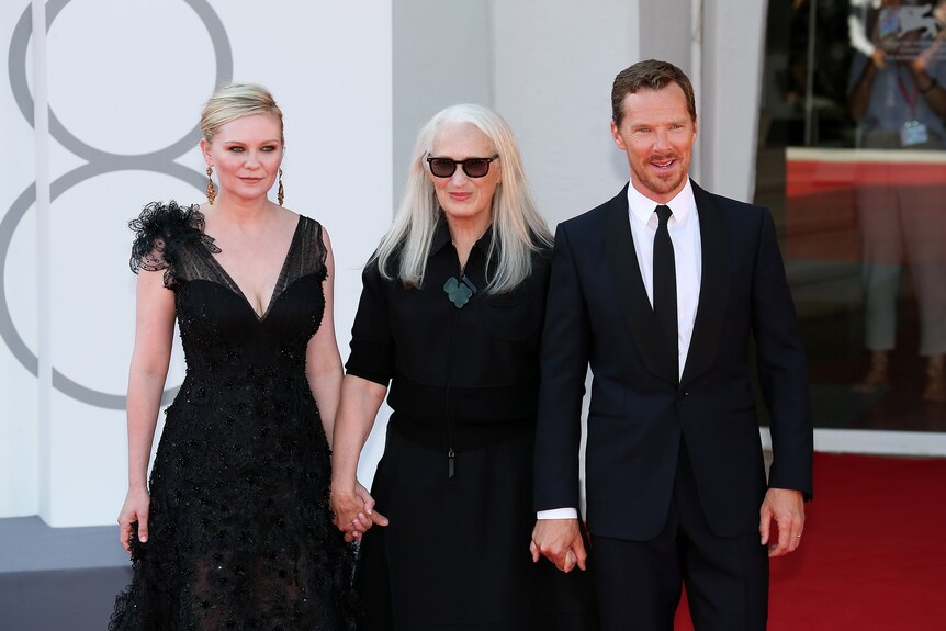 39-year-old blond woman, 67-year-old woman with long white hair and 45-year-old tall white man on red carpet, holding hands.