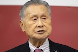 Yoshiro Mori, wearing a black suit, speaks into a microphone