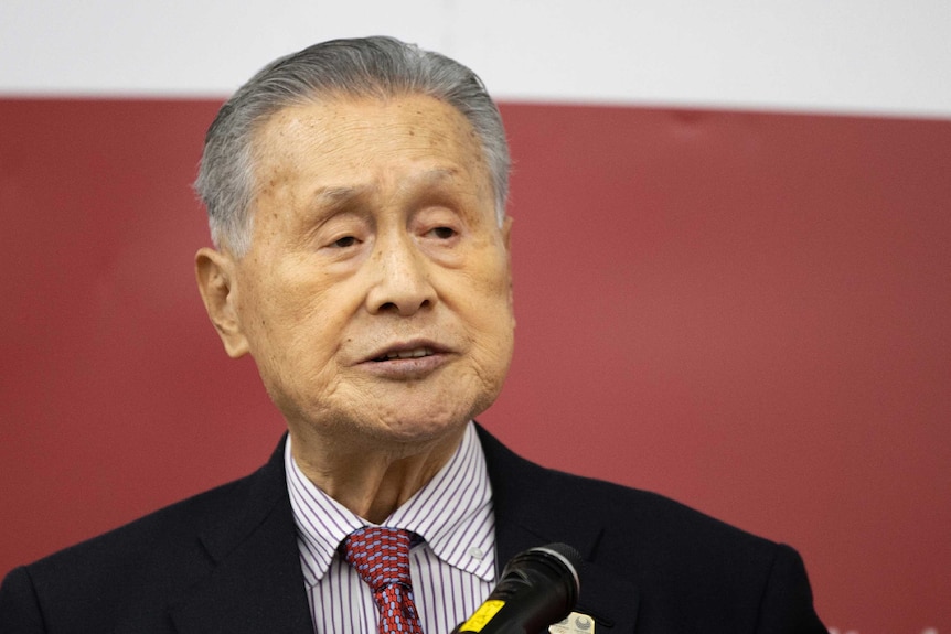 Yoshiro Mori, wearing a black suit, speaks into a microphone