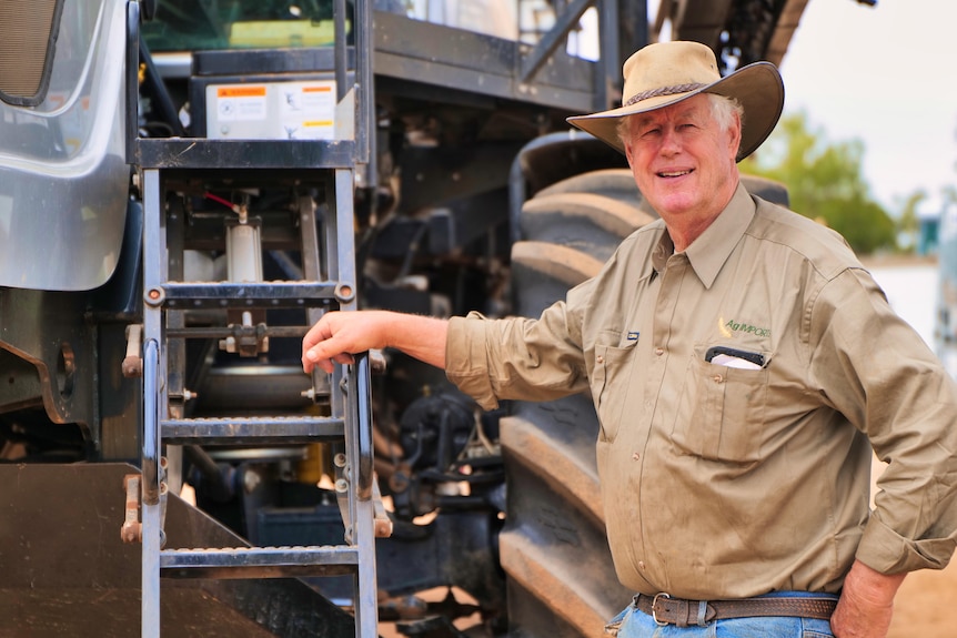 A man in a brown shirt leans on a tractor. He is wearing a full brim hat and is looking at the camera. 