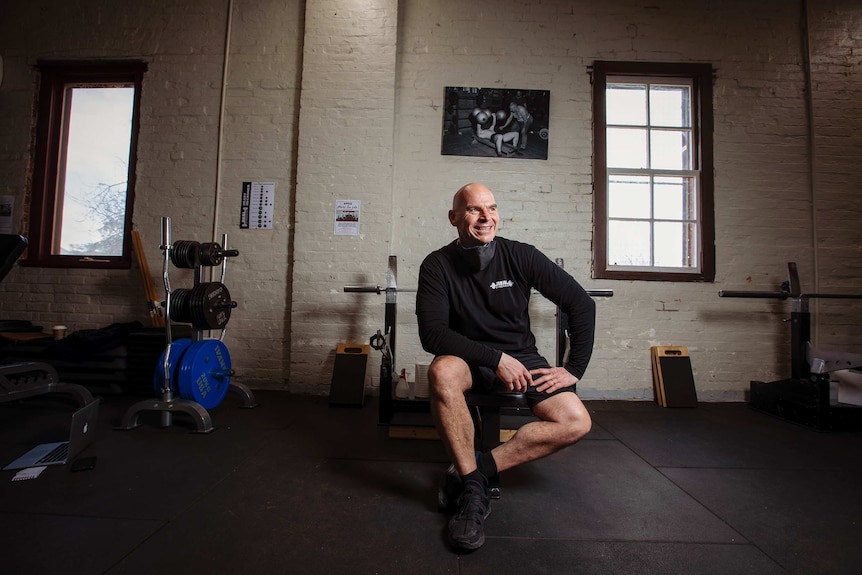 A man in black exercise clothes sits on a stool with gym equipment behind him and a white wall with photograph of weightlifters.