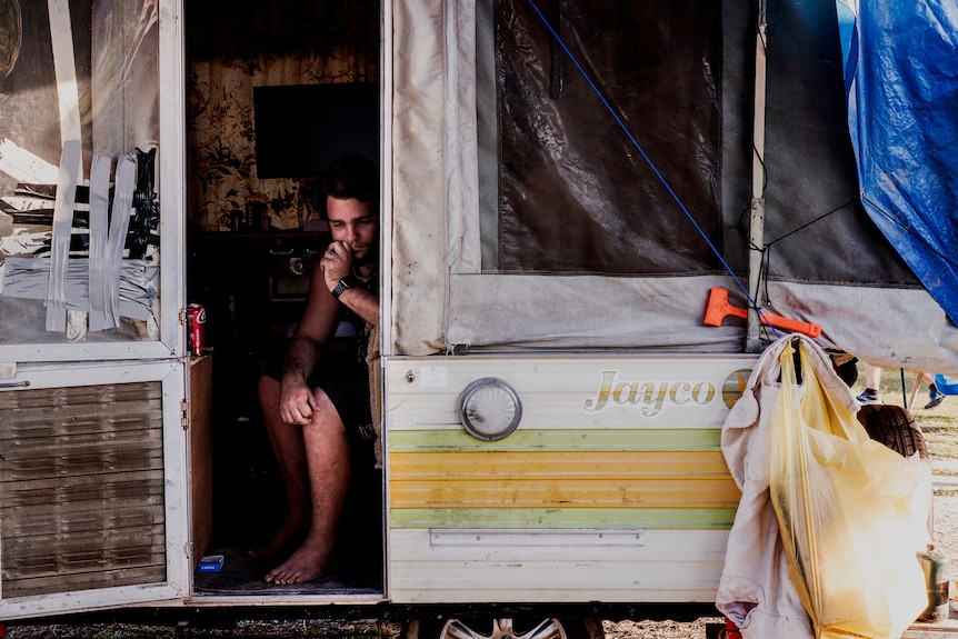 A pensive man young man sits in a rundown caravan door looking down. 