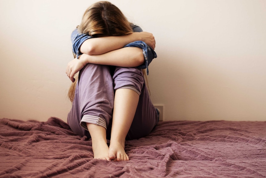 A young woman sits curled up on her bed, her head in her hands.