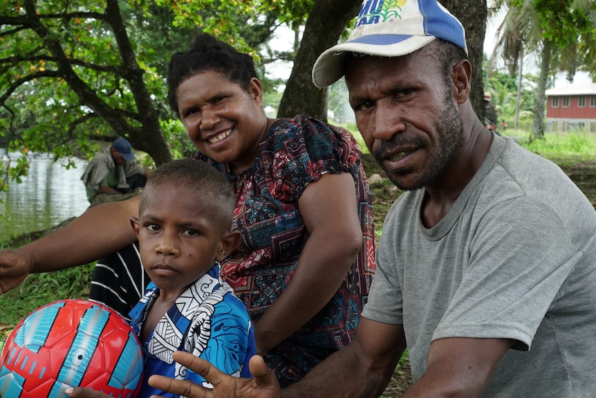 A young boy holding a soccer ball, sitting with his mum and dad in the outdoors.