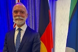 Wiradjuru man wearing a suit and standing in front of the Australian, Aboriginal and Torres Strait Islander flags