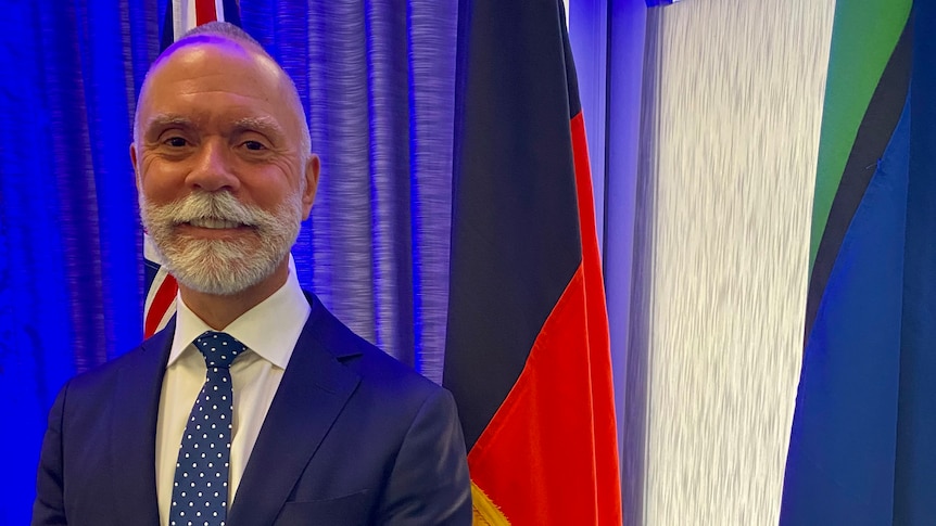 Wiradjuru man wearing a suit and standing in front of the Australian, Aboriginal and Torres Strait Islander flags