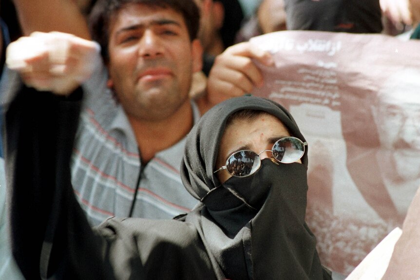 A woman wearing a black jihab and dark glasses raises her fist in a protest crowd