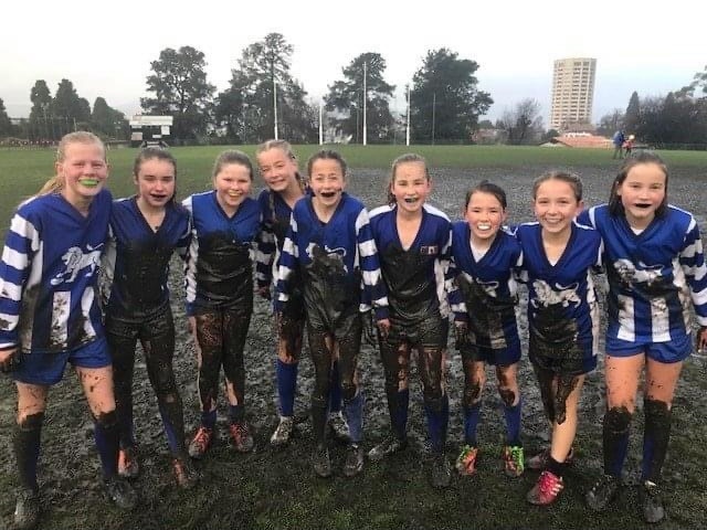 Nine young girls wearing muddy blue and white football jerseys smiling on a football field.