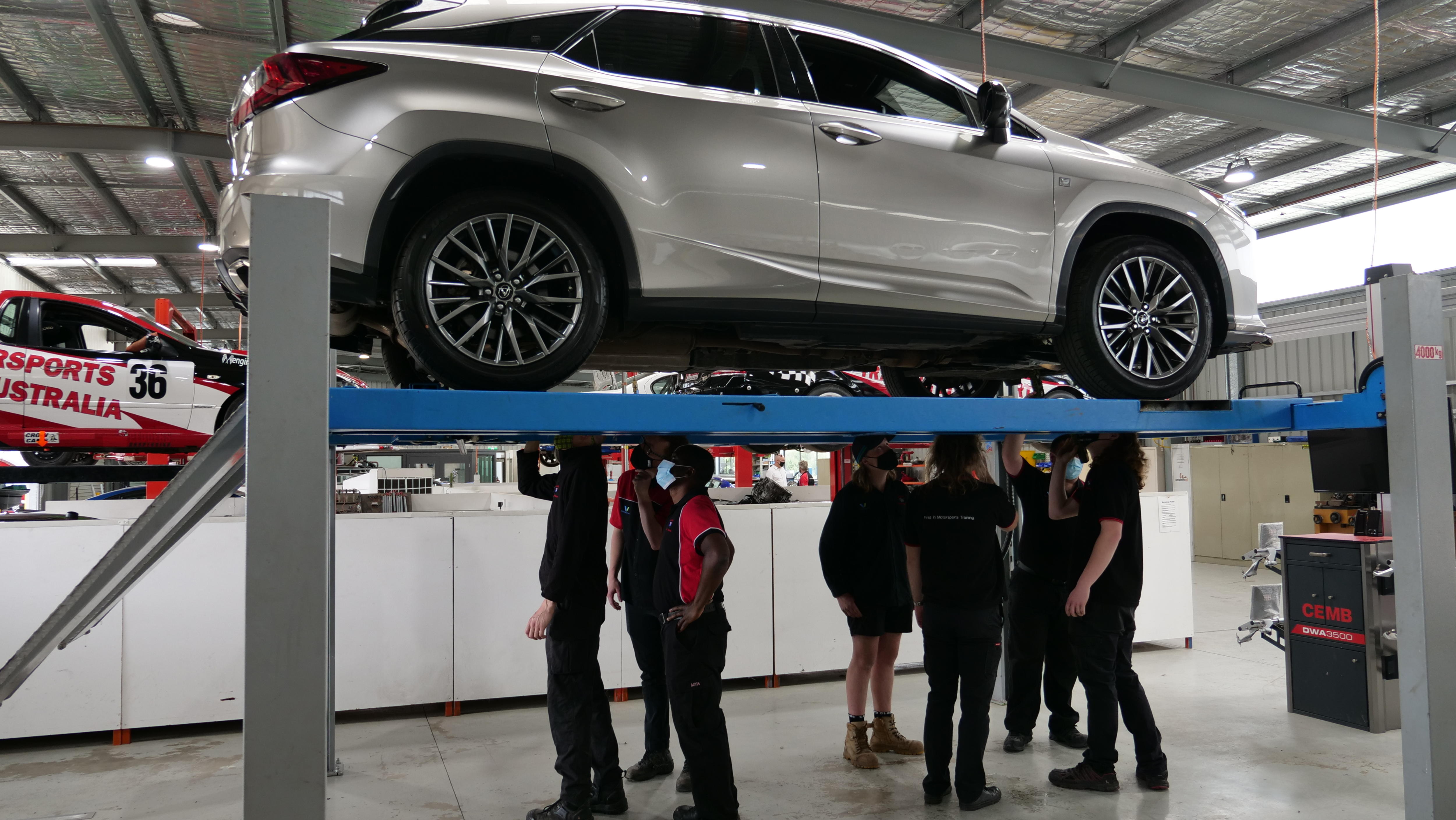 Six people stand underneath a car that is up on a hoist, looking at the chassis. 