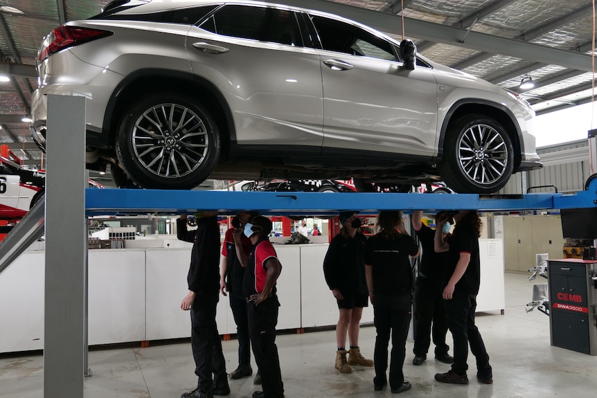 Six people stand underneath a car that is up on a hoist, looking at the chassis. 