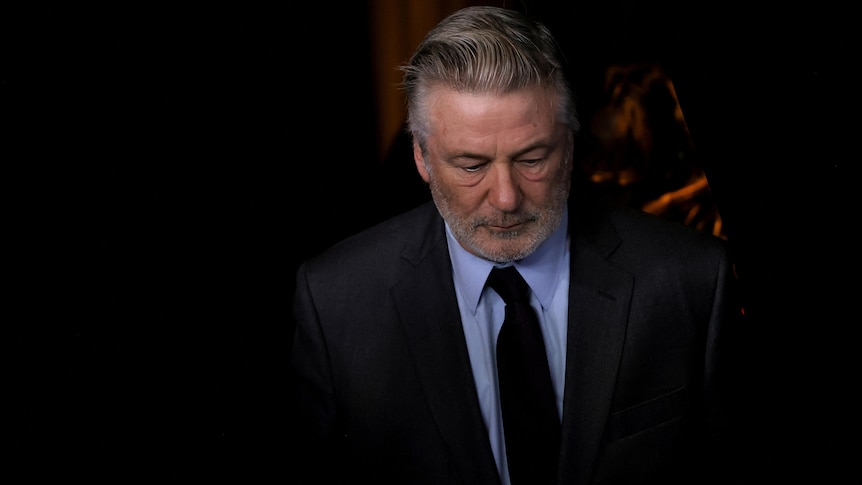 Alec Baldwin looks down wearing a neutral expression, a dark suit and blue shirt