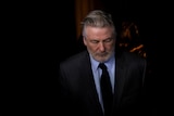 Alec Baldwin looks down wearing a neutral expression, a dark suit and blue shirt