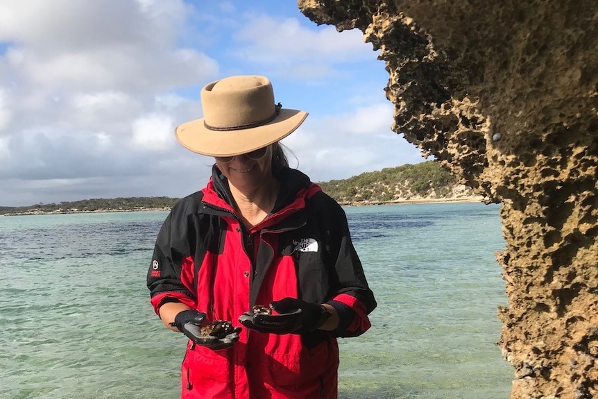 A woman in a red jacket and hat standing in blue ocean water holding micrplastics.