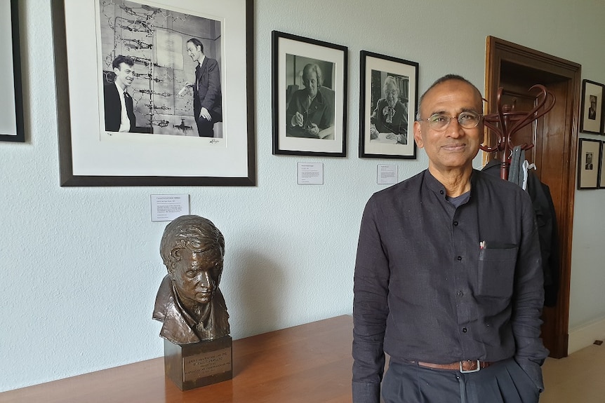 Man in black short and glasses standing next to photos of eminent scientists and a bust of Indian mathematician Ramanujan