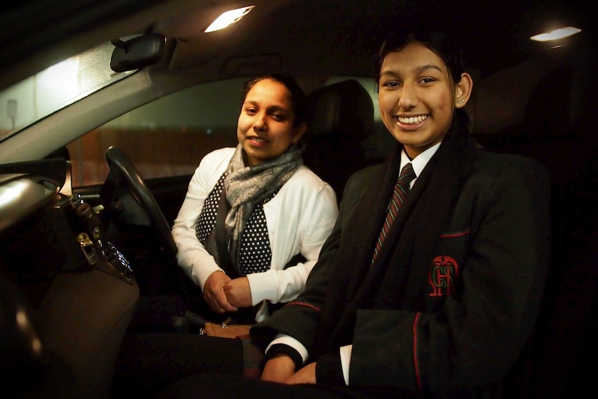 In a car with its roof lights on, you view a mother and her teenage daughter sitting and smiling into the camera.