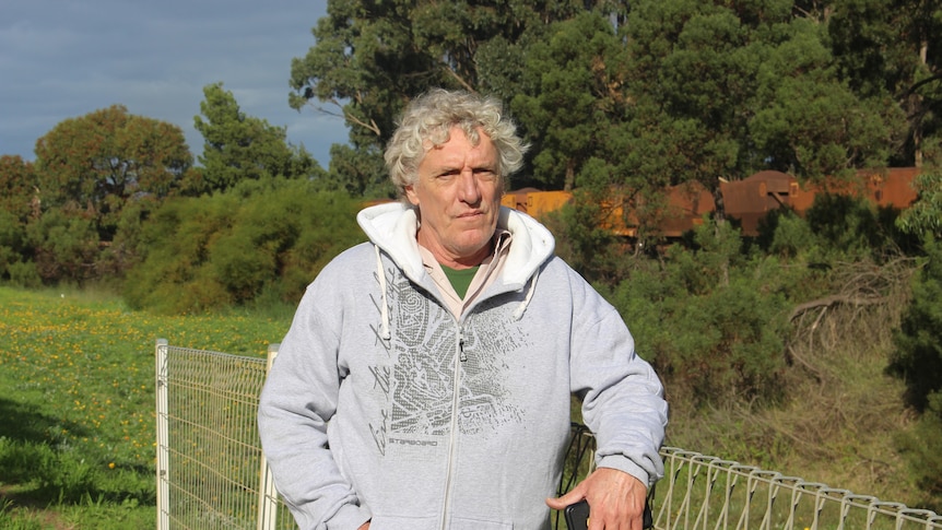 A silver-haired man stands in a hoodie as a train passes behind him.