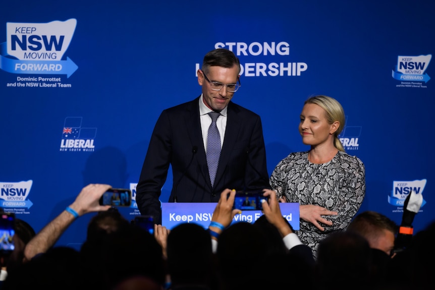 Dominic Perrottet stands behind a lectern on NSW election with his wife Helen Perrottet