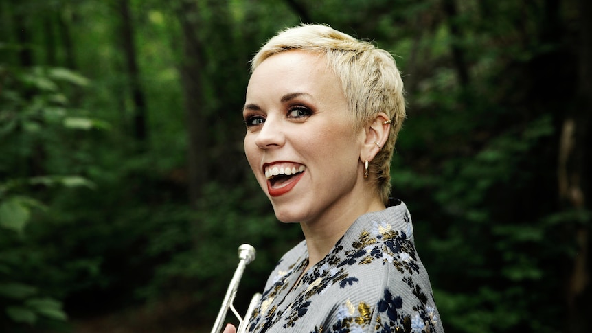 A woman stand side on looking at the camera. She is smiling and holding a trumpet.