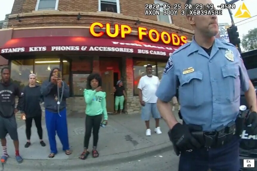 Derek Chauvin stands outside a shop in Minneapolis in front of a number of pedestrians in a still from police body cam