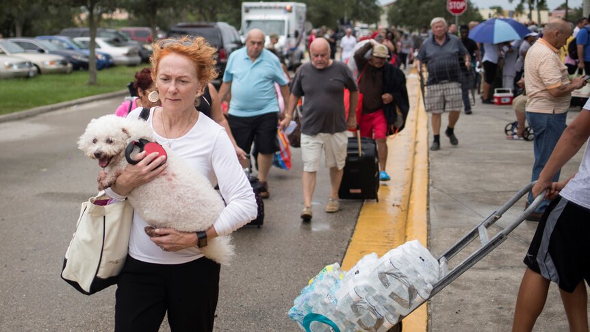 A woman holds a small white dog to her chest as her and other residents walk down a street carrying their belongings.