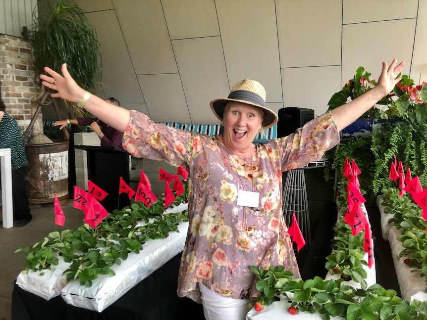 A woman wearing a hat smiles and poses with numbered strawberry plants around her.