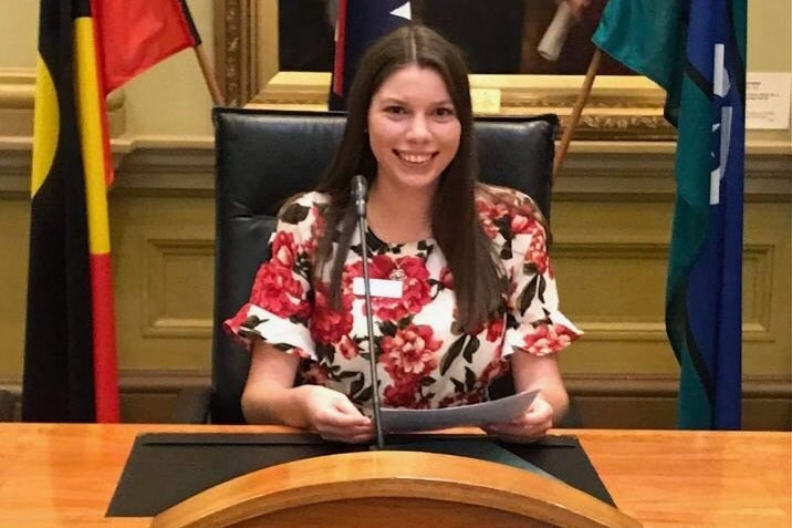 Une femme aux cheveux bruns, vêtue d'un chemisier blanc et rouge, est assise à une table du conseil dans la salle du conseil.