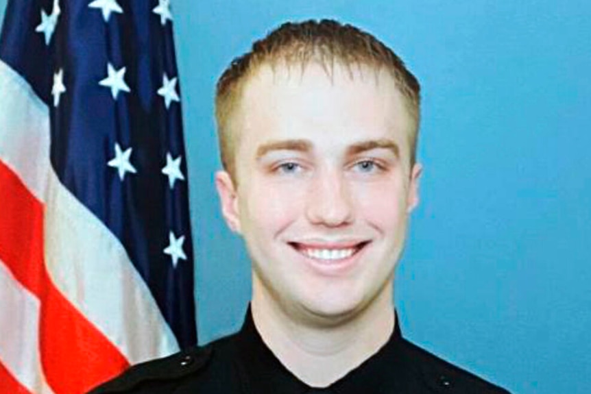 Blonde-haired man in black uniform in front of American flag