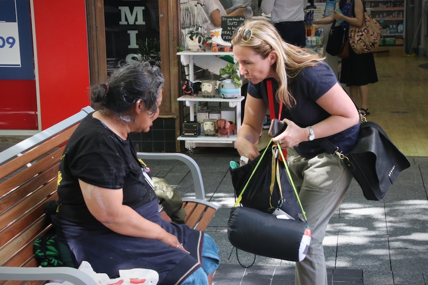 A woman bends down passing bags and supplies to another woman sitting on a bench