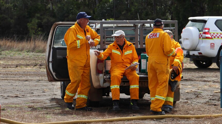 Four firefighters in CFA protective gear sit and stand near a ute, talking.