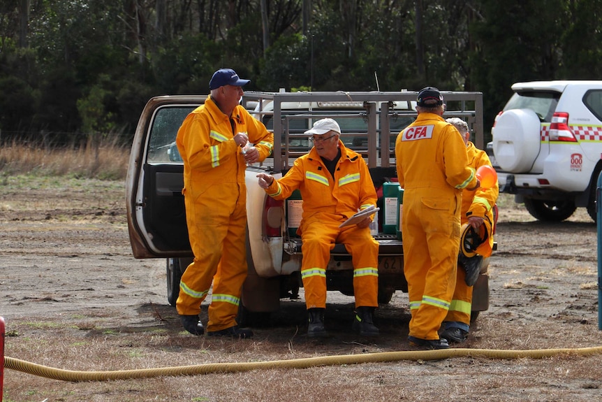 Four firefighters in CFA protective gear sit and stand near a ute, talking.