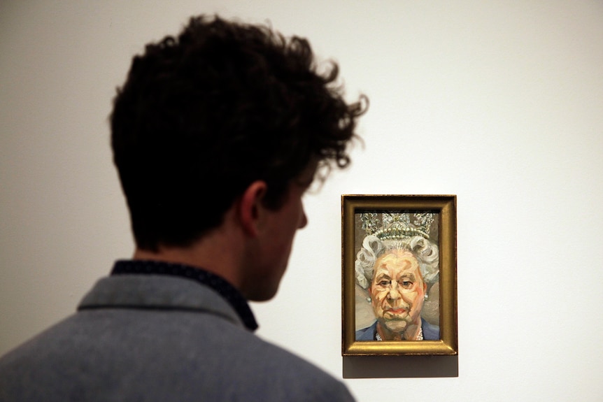 The back of a man's head fills most of the frame as he looks at a small, wall-mounted portrait of a scowing Queen Elizabeth II.