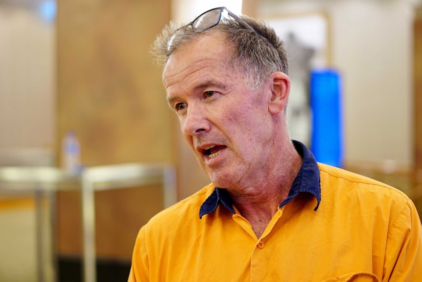 Mike Hollier in mid-speech, wearing a yellow shirt and reading glasses on his forehead.