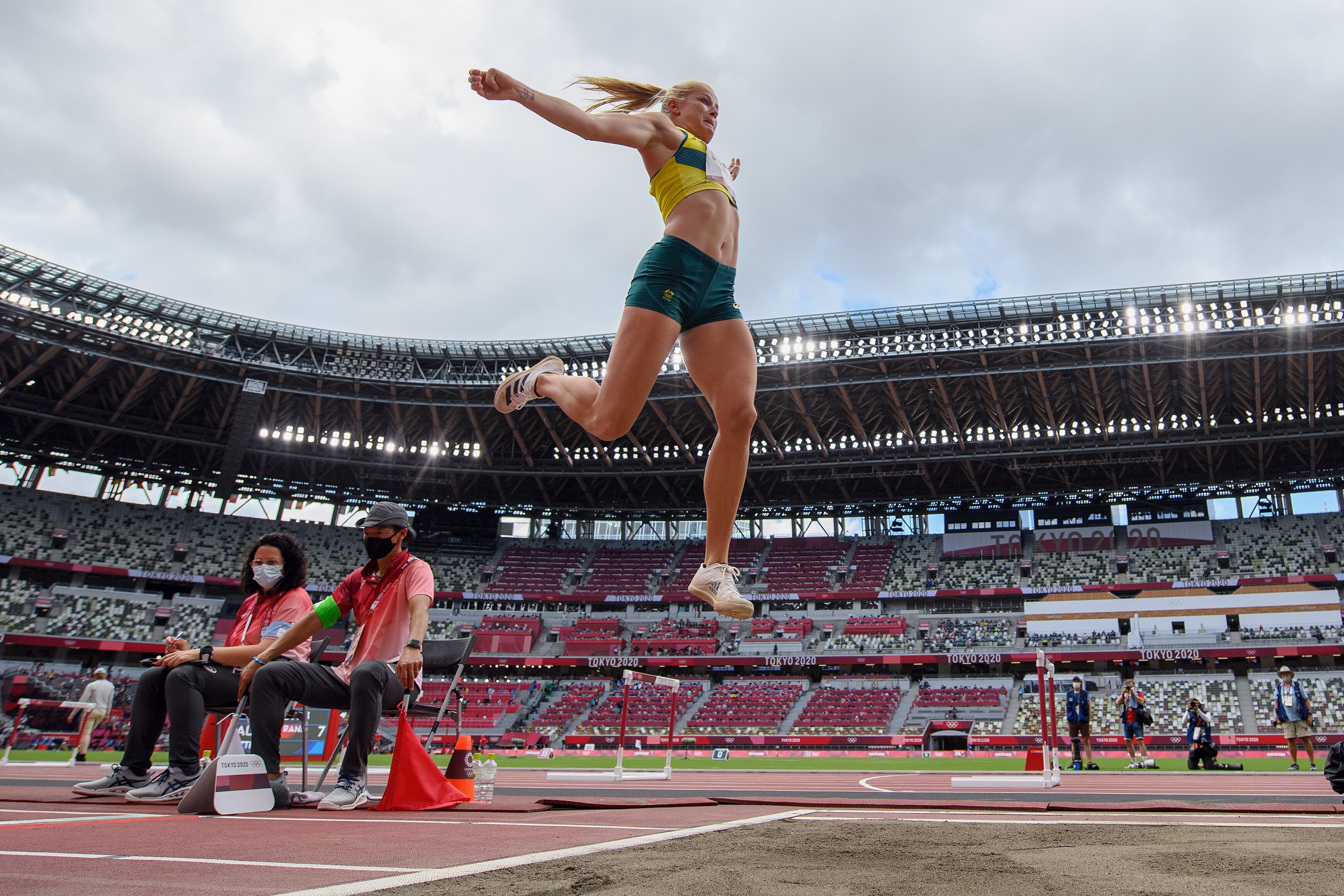 A woman in green and gold jumps high in the air above a sand pit