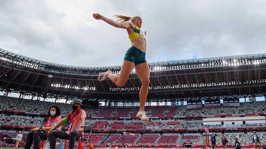 A woman in green and gold jumps high in the air above a sand pit