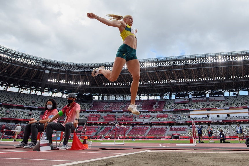A woman in green and gold jumps high in the air above a sand pit