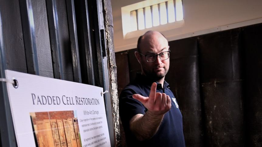 A bespectacled man stands outside a padded cell, beckoning with his finger and a sinister expression on his face.