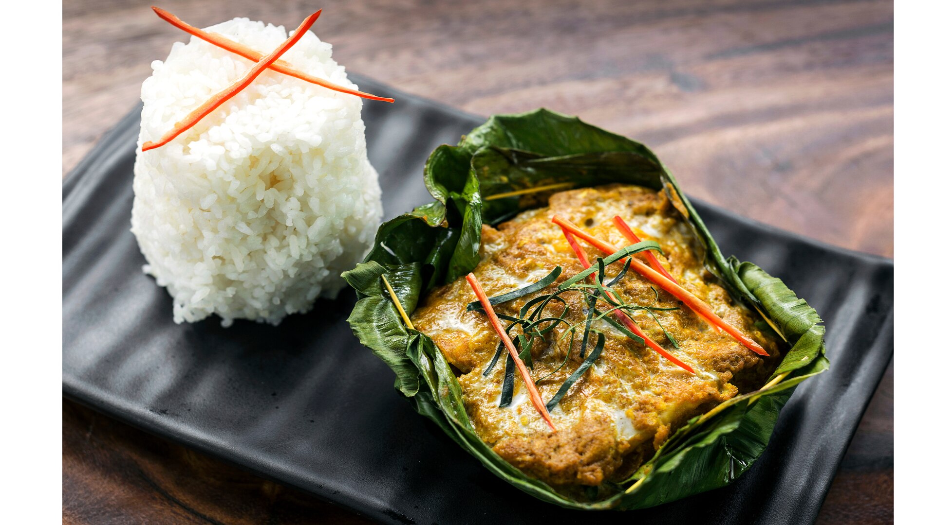 A serving of fish amok wrapped in banana leaves with a side of white rice on a black plate.