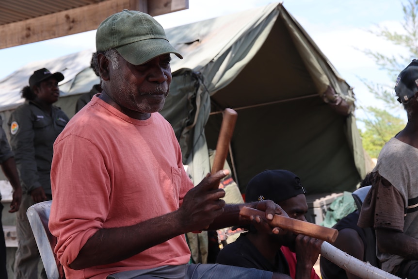 An older Indigenous man wearing a hat uses clapping sticks.
