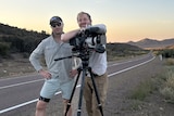 Two men stand behind a video camera next to a highway. It's dusk and a mountain range is lit up in the background