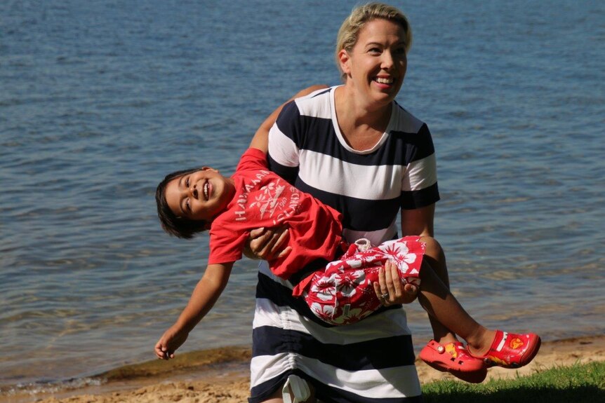 A woman holds a young boy in a red t-shirt and shorts while walking on the beach.