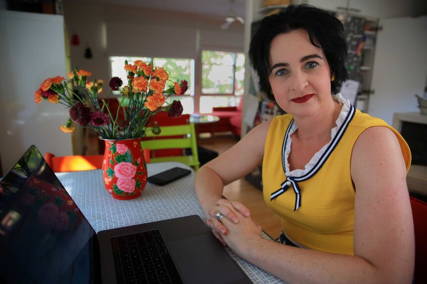 A woman looks into the camera while sitting at a table with a laptop on it, a vase of flowers in the background.