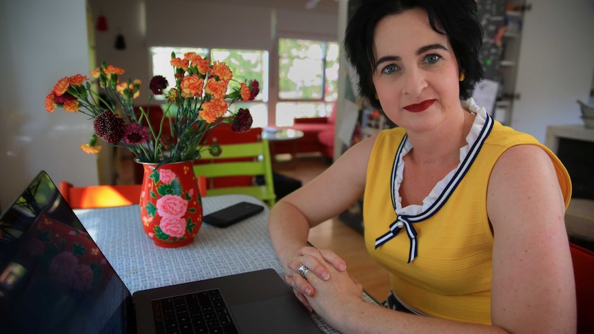A woman looks into the camera while sitting at a table with a laptop on it, a vase of flowers in the background.
